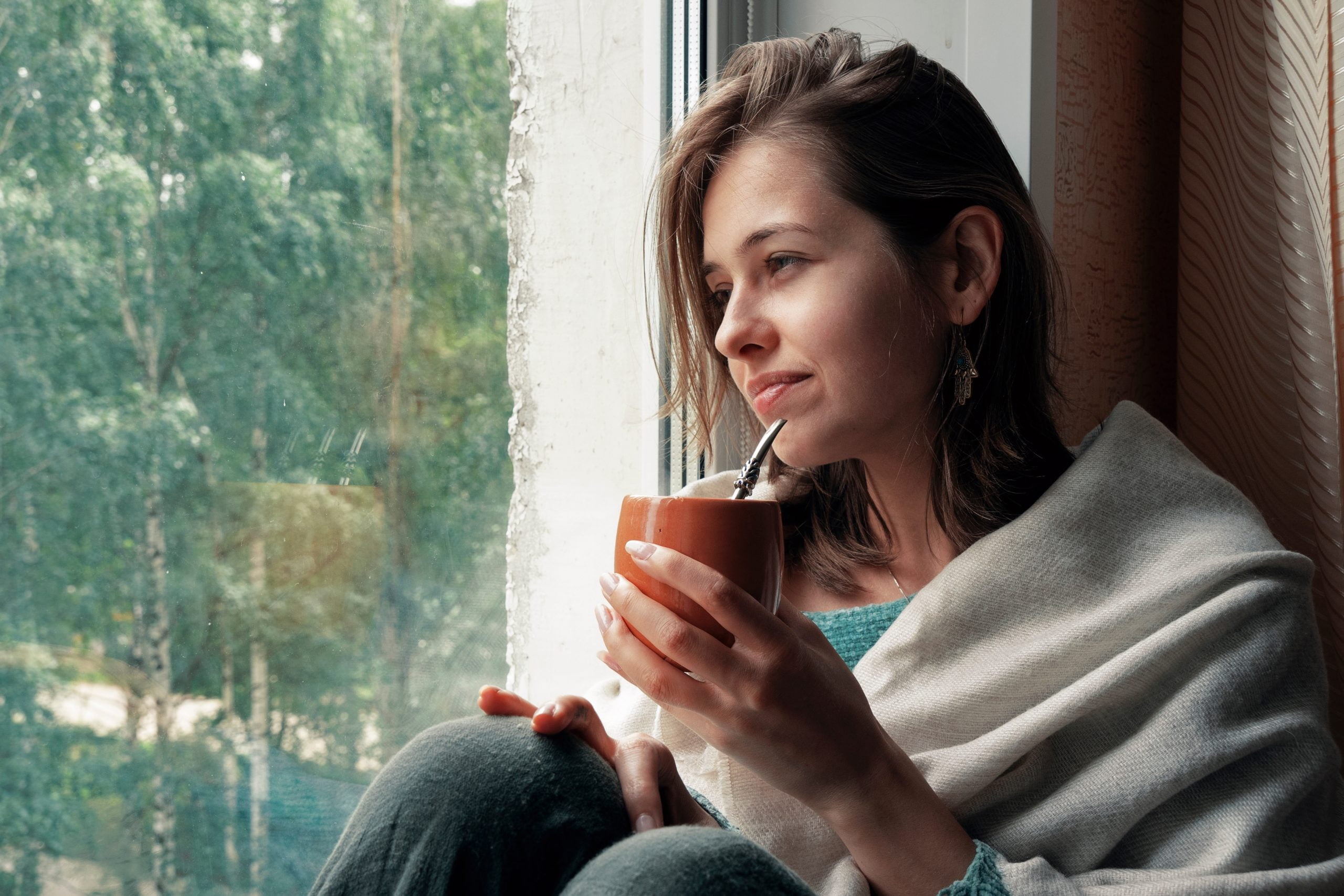 Young woman drinks mate on the windowsill by the window. Concept of mental health